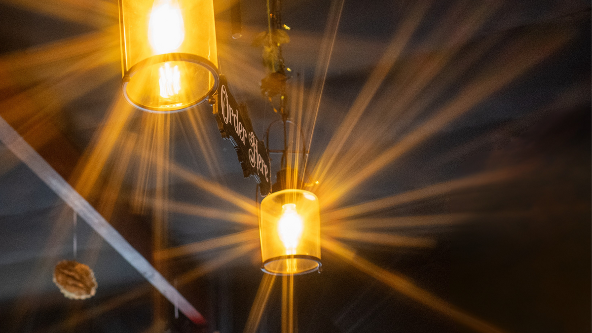 Two warm yellow hanging pendant lights with glowing bulbs, shown in a dark interior setting with dramatic light rays around the Order Here sign hanging from the ceiling.